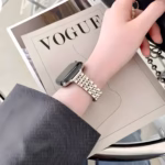 Bracelet Apple Watch EzStrap en acier inoxydable au design lisse et raffiné, style luxe, porté au poignet d’une personne lisant un magazine sur une table en verre.