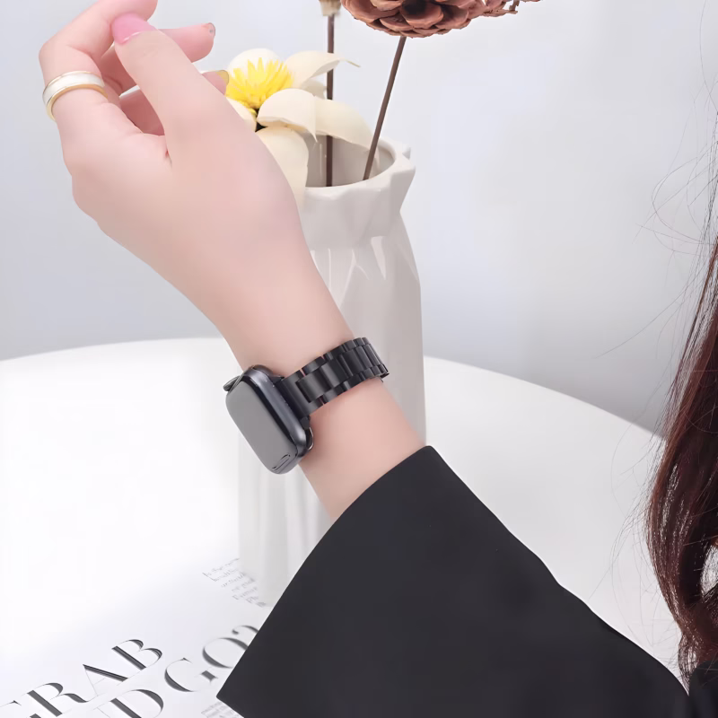 Bracelet Apple Watch EzStrap en métal noir, à maillons classiques au style élégant, porté au poignet d’une femme en tenue habillée, avec un vase de fleurs en arrière-plan.