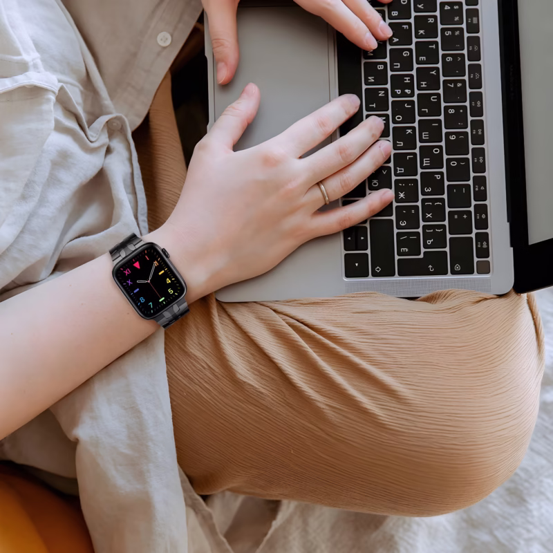 Bracelet Apple Watch EzStrap en acier inoxydable lisse, style luxe, avec maillons angulaires, porté par une personne travaillant sur un ordinateur portable.