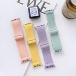 Bracelets Apple Watch EzStrap en nylon souple texturé de style sport, déclinés en coloris pastel (rose, jaune, lavande et vert), posés à plat sur une table blanche à côté d'une bougie et d'un cadran de montre.