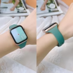 Bracelet Apple Watch EzStrap en silicone lisse vert, au style sport, porté au poignet d’une femme sur un fond en dentelle avec un magazine en arrière-plan.