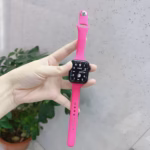 Bracelet Apple Watch EzStrap en silicone rose lisse de style sport, tenu à la main devant un mur clair avec une plante en arrière-plan.
