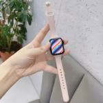 Bracelet Apple Watch EzStrap en silicone lisse rose pâle, de style sport, tenu en main devant un mur texturé et un canapé gris.