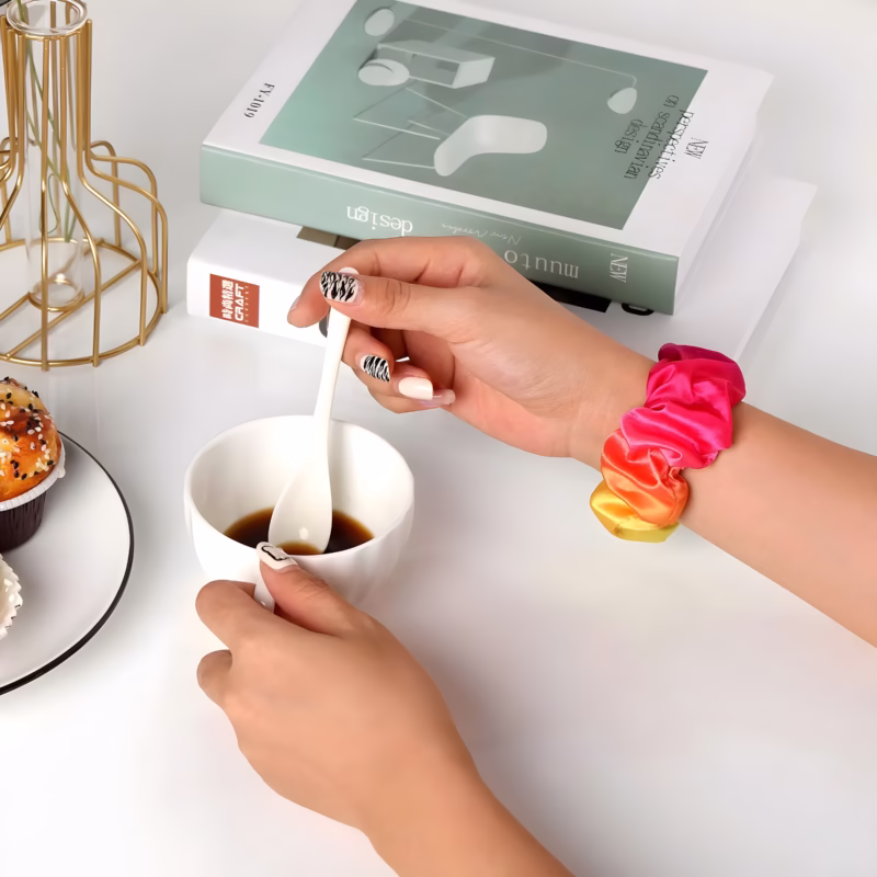 Bracelet Apple Watch EzStrap en textile élastique style chouchou, aux tons dégradés rose, orange et jaune, porté au poignet d'une femme prenant un café à une table blanche.