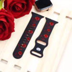 Bracelet Apple Watch EzStrap en silicone élégant noir, gravé d’un motif floral rouge, posé sur une planche en bois à côté de roses rouges.