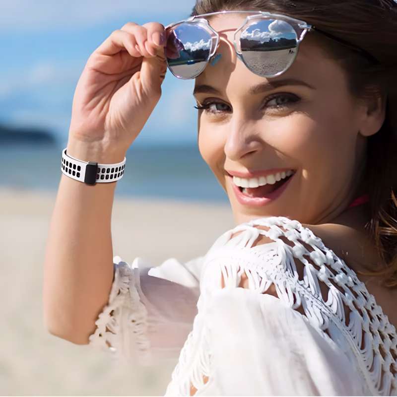 Bracelet Apple Watch EzStrap en silicone perforé blanc et noir, au style sport, porté au poignet d’une femme souriante sur une plage ensoleillée.