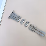 Bracelet Apple Watch EzStrap en métal inoxydable à construction articulée, style luxe avec finition lisse, posé en pièces détachées sur une surface beige.