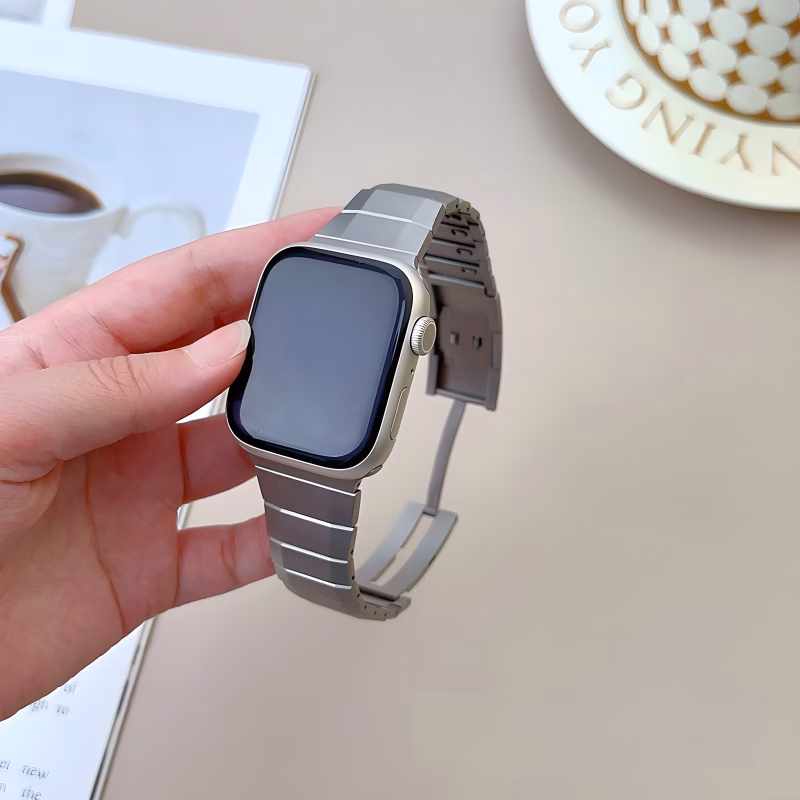Bracelet Apple Watch EzStrap en acier inoxydable lisse, style premium, tenu en main au-dessus d'une table beige avec une tasse de café en arrière-plan.