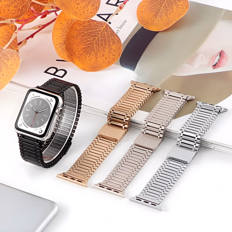 Bracelet Apple Watch EzStrap en acier inoxydable à maille articulée lisse, style luxe, présenté en quatre coloris métalliques posés sur un magazine de mode avec des feuilles décoratives en arrière-plan.
