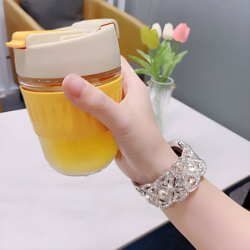 Bracelet Apple Watch EzStrap en acier inoxydable de style luxe, orné de maillons décoratifs sertis de cristaux scintillants, porté au poignet d’une femme tenant un mug jaune dans un intérieur moderne.