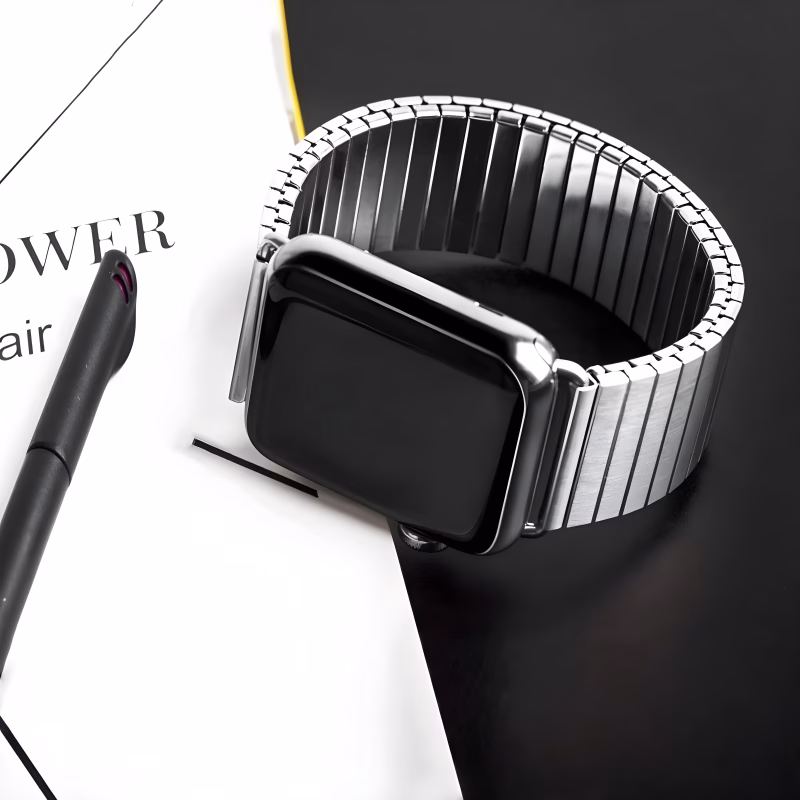 Bracelet Apple Watch EzStrap en métal brossé, élégant et à maillons flexibles extensibles, posé sur une table noire à côté d’un stylo et d’un carnet.