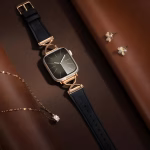 Bracelet Apple Watch EzStrap en cuir noir grainé avec fixation en métal doré ornée en X, de style luxe, posé sur un fond en cuir brun avec bijoux en arrière-plan.