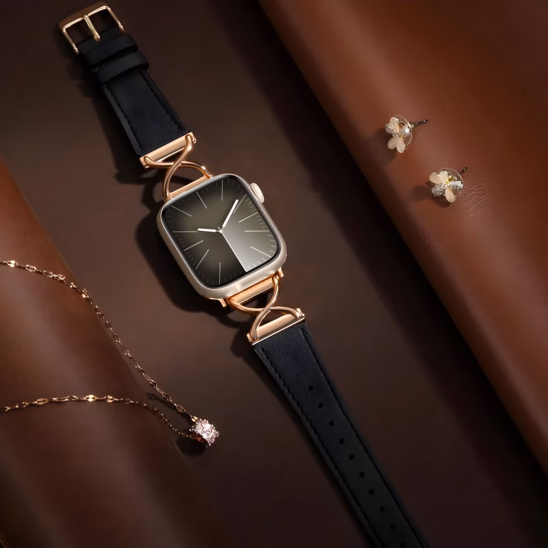 Bracelet Apple Watch EzStrap en cuir noir grainé avec fixation en métal doré ornée en X, de style luxe, posé sur un fond en cuir brun avec bijoux en arrière-plan.