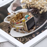 Bracelet Apple Watch EzStrap en cuir multibrins avec ornements métalliques, au style artisanal et élégant, incluant des motifs colorés et des touches scintillantes, posé sur un magazine illustrant une tasse de café.