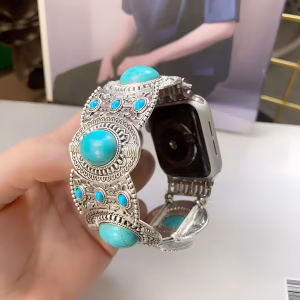 Bracelet Apple Watch EzStrap en métal alliage de zinc, style élégant et artisanal, orné de pierres turquoises serties avec motifs gravés et texture ciselée, tenu en main devant une personne assise en arrière-plan.