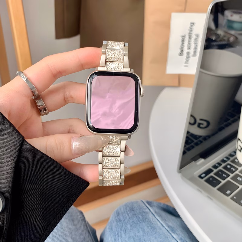 Bracelet Apple Watch EzStrap en métal alliage de zinc, style premium brillant avec structure articulée décorée de strass scintillants, tenu à la main par une personne en veste noire devant un ordinateur portable.