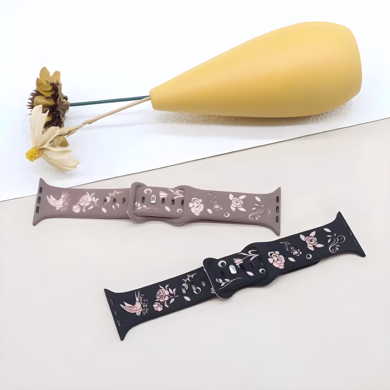 Bracelet Apple Watch EzStrap en silicone doux, style artisanal, décor gravé floral et oiseau stylisé, posé à plat sur une surface beige à côté d’un vase jaune et de fleurs séchées.