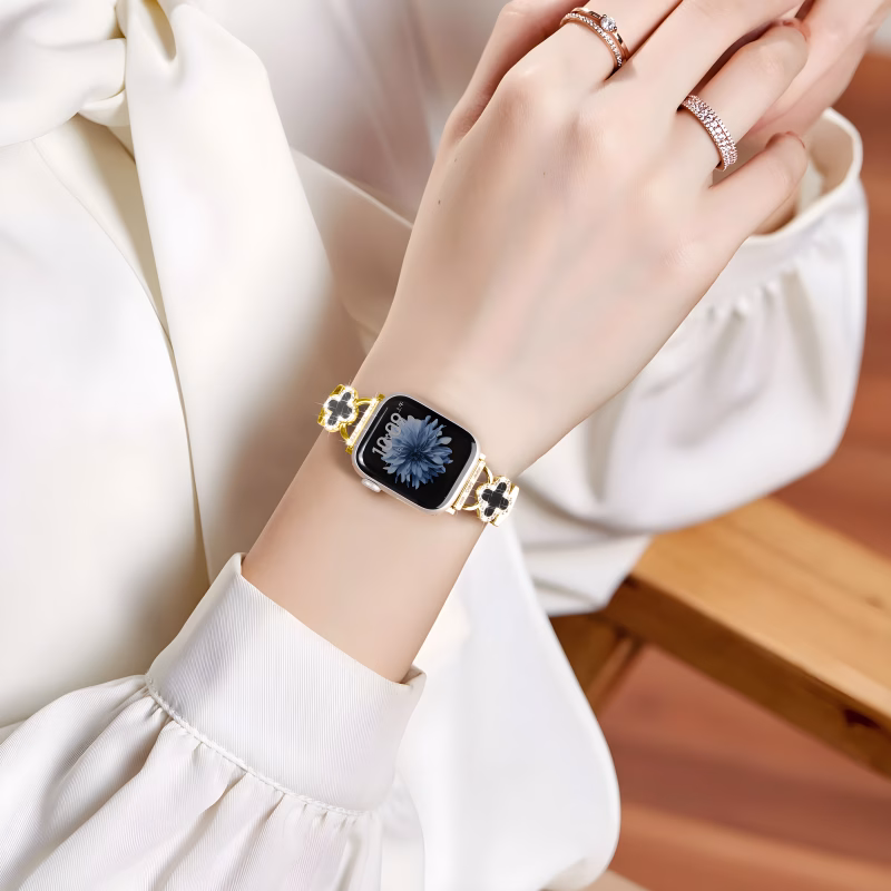 Bracelet Apple Watch EzStrap en métal brillant de style luxe, orné de trèfles à cristaux noirs, porté au poignet d'une femme en blouse blanche, assise sur une chaise en bois.