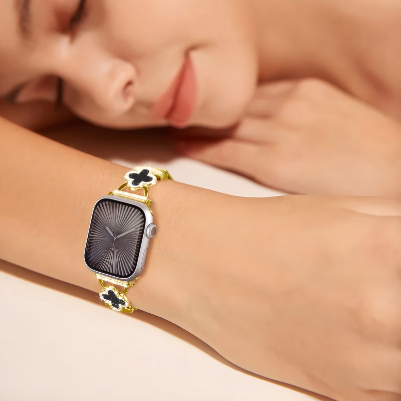 Bracelet Apple Watch EzStrap en métal doré style milanais, orné de trèfles à cristaux noirs brillants, porté au poignet d'une femme allongée.