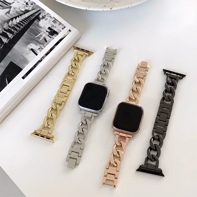 Bracelet Apple Watch EzStrap en métal lisse, style luxe, avec design de maillons géométriques et ovales, présenté en quatre coloris posés à plat sur une table blanche à côté d’un magazine et d’une assiette.