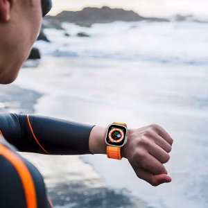 Bracelet Apple Watch EzStrap en métal avec structure tissée de style sport, motif ondulé orange, porté au poignet d’un homme en tenue de plongée au bord de l’océan.