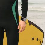 Bracelet Apple Watch EzStrap en nylon tressé noir, de style sport avec texture texturée, porté au poignet d’un homme en combinaison de surf tenant une planche au bord de la mer.