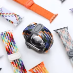 Bracelet Apple Watch EzStrap en plastique tressé de style sport, avec un design multicolore texturé, posé en gros plan sur une surface blanche aux côtés d'autres bracelets similaires.