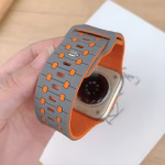 Bracelet Apple Watch EzStrap en silicone perforé bicolore gris et orange, de style sportif, tenu en gros plan dans une main au-dessus d'une montre posée sur un carnet.