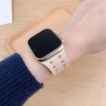 Bracelet Apple Watch EzStrap en silicone blanc perforé, de style sport, avec découpes orange, porté au poignet d’une personne sur fond de table en bois clair.