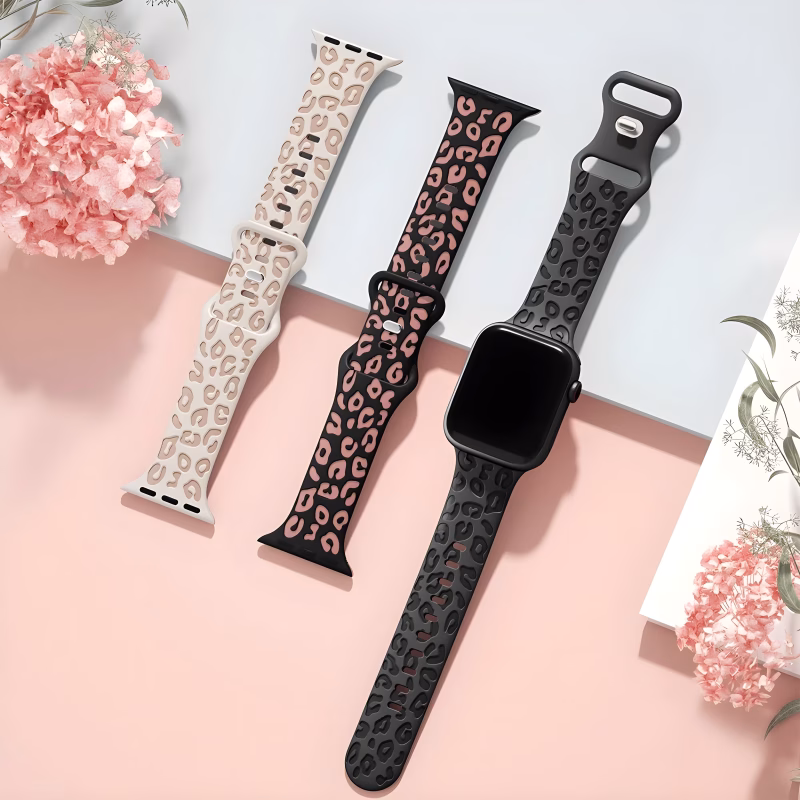 Bracelet Apple Watch EzStrap en silicone gravé à motif léopard, décliné en trois coloris, posé à plat sur une surface rose pastel parmi des fleurs décoratives.