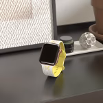 Bracelet Apple Watch EzStrap en silicone élégant, de couleur blanche et jaune, avec motif floral découpé, présenté debout sur une table noire près d’une bougie décorative.