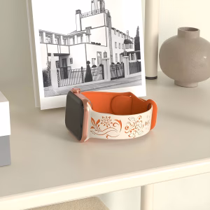 Bracelet Apple Watch EzStrap en silicone élégant, blanc et orange, à motif floral gravé, posé sur une table beige près d’un vase et d’un livre.