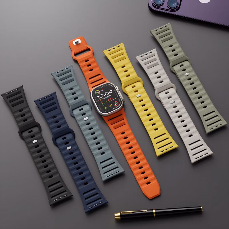Bracelets Apple Watch EzStrap en silicone mat perforé, au style sport, déclinés en plusieurs couleurs, disposés à plat sur une surface noire à côté d’un stylo et d’un iPhone.