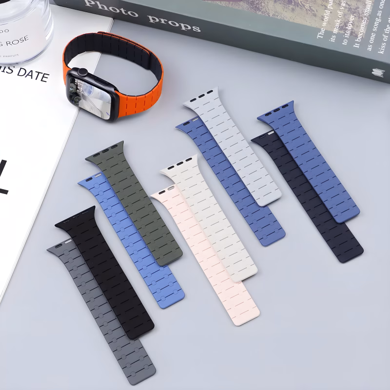 Bracelet Apple Watch EzStrap en silicone texturé de style sport, décliné en plusieurs coloris, posé à plat sur une surface grise, avec une montre équipée d’un bracelet orange en arrière-plan.