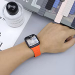 Bracelet Apple Watch EzStrap en silicone texturé de style sport, de couleur orange vif, porté au poignet d’un homme sur un bureau avec un flacon de parfum et des bracelets en arrière-plan.
