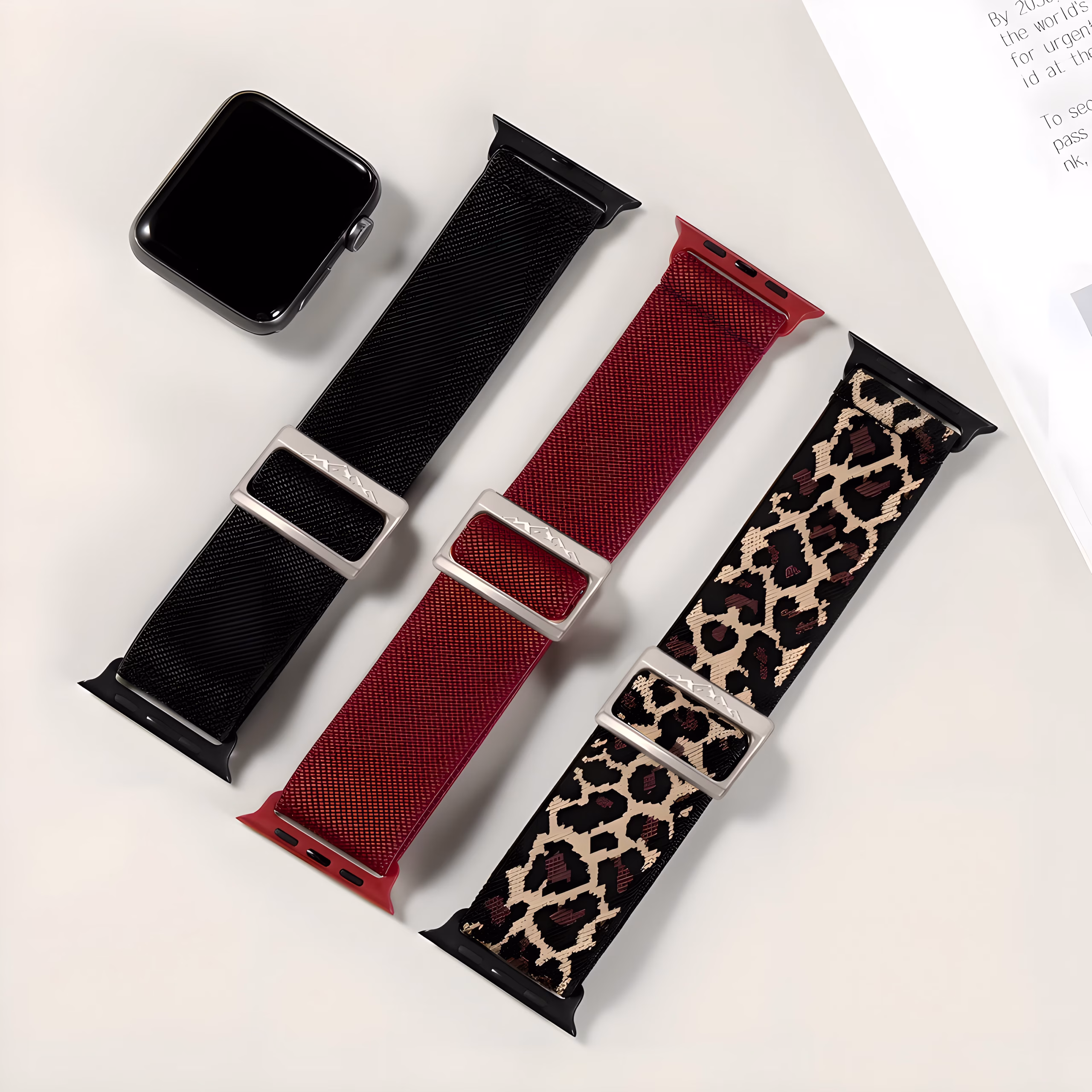 Bracelet Apple Watch EzStrap en nylon texturé, style sport, décliné en noir uni, rouge vif, et motif léopard, posés sur une surface blanche à côté d’une montre et d’un magazine.