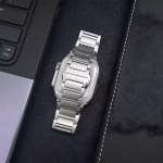 Bracelet Apple Watch EzStrap en titane brossé, style luxe à maillons articulés, présenté posé à plat entre un ordinateur portable et un étui en cuir.