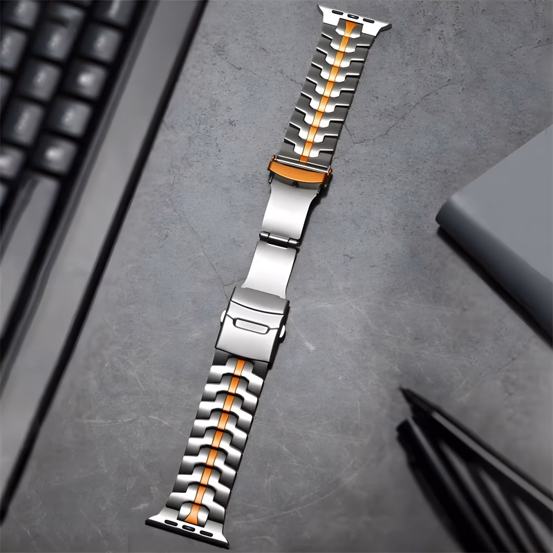 Bracelet Apple Watch EzStrap en métal argenté, style moderne avec bande orange centrale, posé sur un bureau en béton aux côtés d’un clavier et d’accessoires de papeterie.