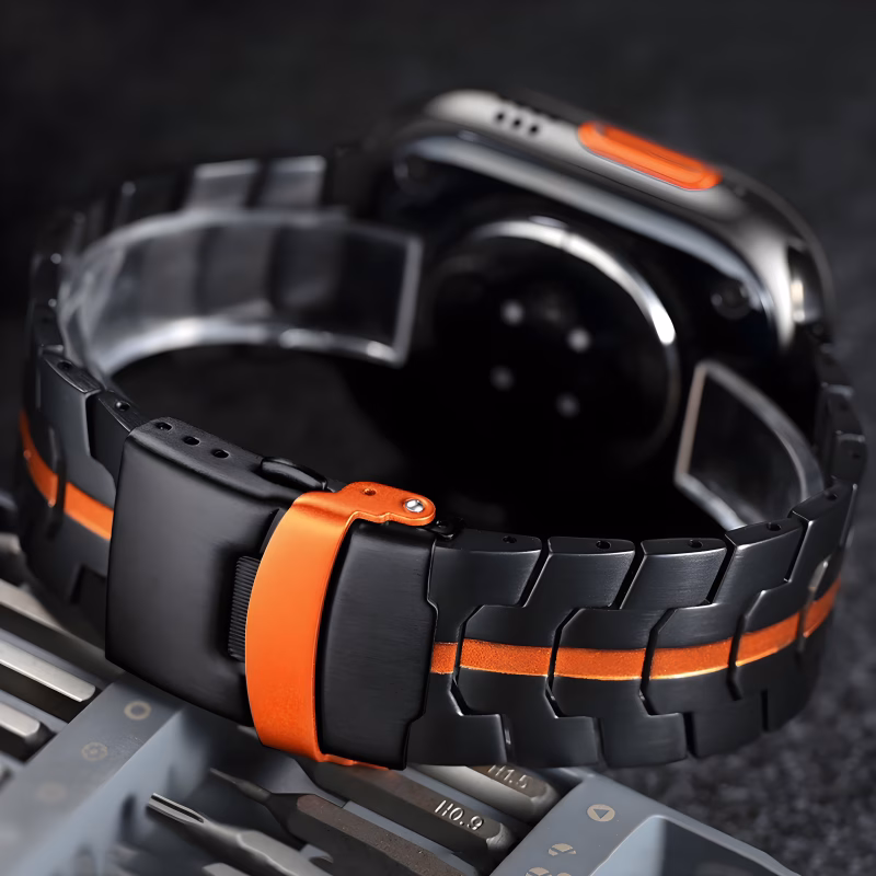 Bracelet Apple Watch EzStrap en métal noir avec bande orange centrale de style sport, présenté en gros plan sur une surface sombre aux côtés d’outils métalliques.